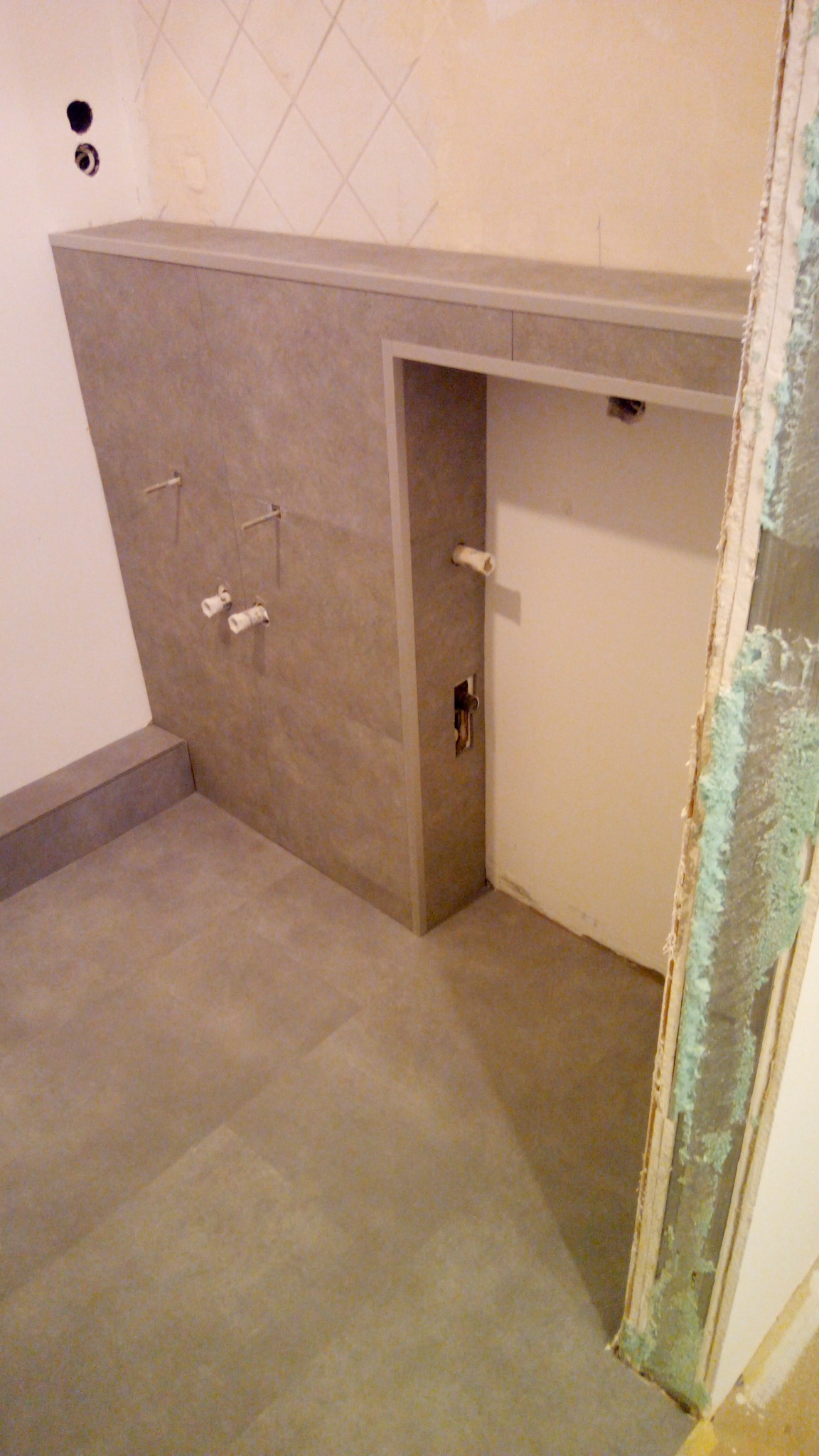 Szara, matowa glazura ścienna i podłogowa w trakcie montażu w nowej łazience, widoczne wystające rury i kable instalacji wodnej i elektrycznej, surowa ściana z pozostałościami po starej glazurze...