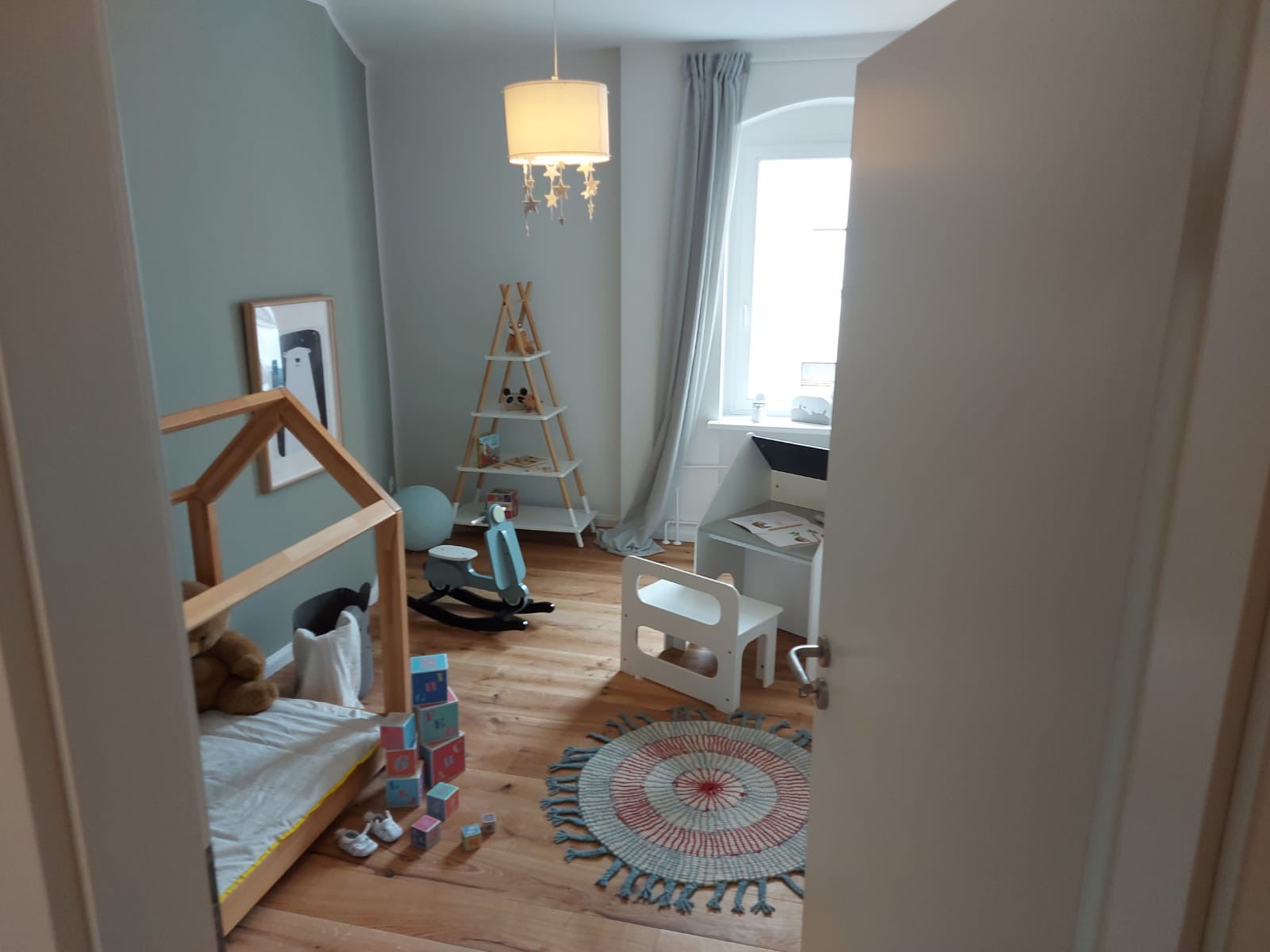 Jasny pokój dziecięcy z drewnianą podłogą, łóżkiem w kształcie domku, biurkiem przy oknie i pastelowymi ścianami, widziany zza uchylonych drzwi.