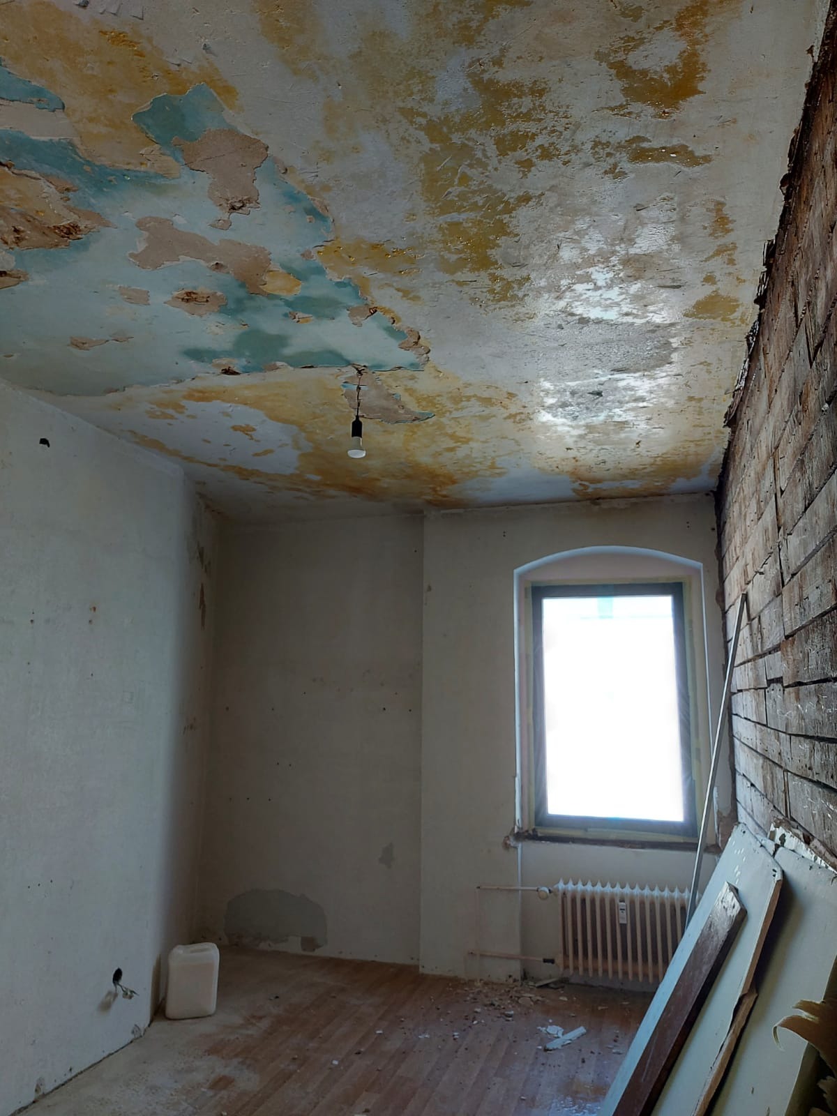 Pokój w trakcie prac remontowych: zniszczony sufit z odpadającą farbą, fragment ściany z odsłoniętą cegłą, okno z grzejnikiem, podłoga z resztkami gruzu, plastikowy kanister przy ścianie.