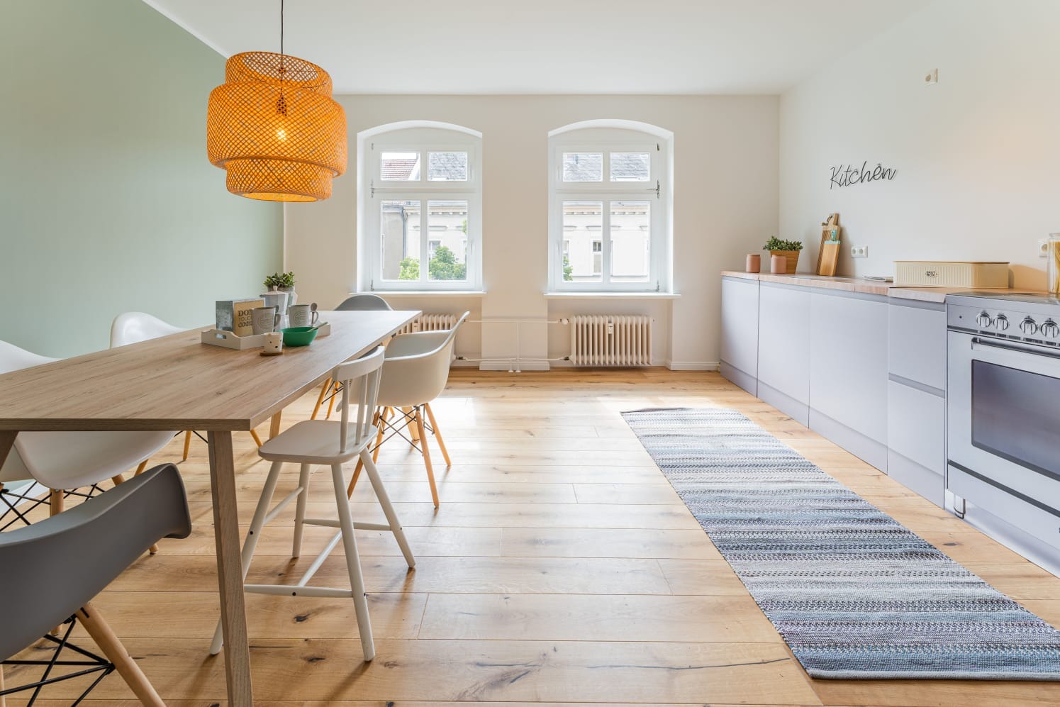 Jasne wnętrze z drewnianą podłogą, stołem z krzesłami w stylu skandynawskim, szarą zabudową kuchenną i plecionym dywanem w odcieniach szarości i błękitu.