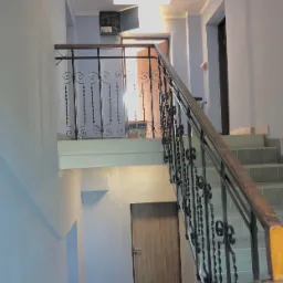 Klatka schodowa w budynku, ściany pomalowane na jasny kolor, widoczne schody z szarymi płytkami, ozdobna metalowa balustrada i drzwi wejściowe. Na podeście widoczne wiadro z farbą.