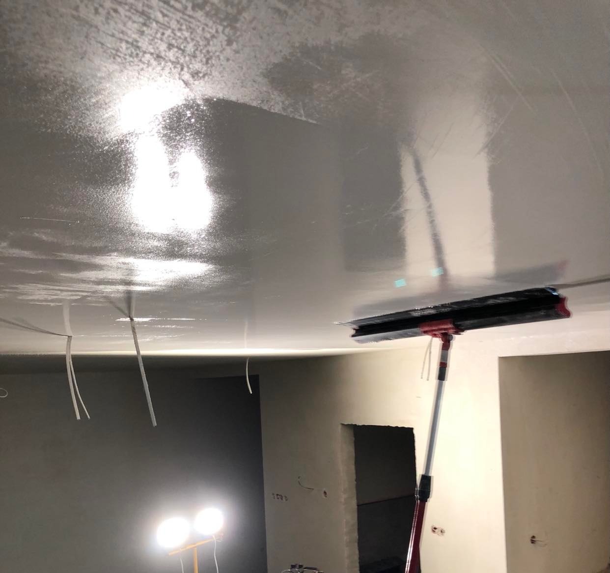 Aplikacja mokrej gładzi na suficie za pomocą specjalnego aplikatora na teleskopowym wysięgniku, widoczne przewody elektryczne i lampa budowlana.