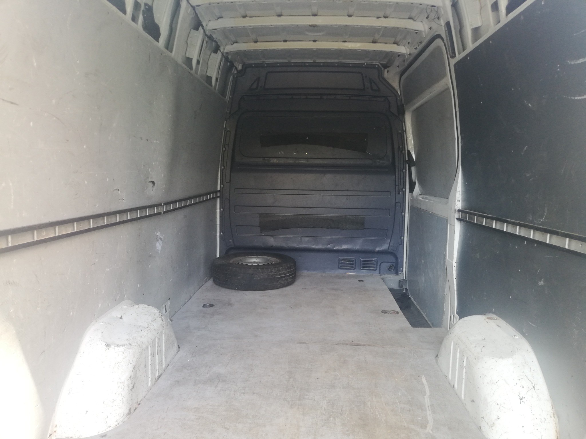 Pusta przestrzeń ładunkowa białego furgonetki z otwartymi tylnymi drzwiami, widoczne nadkole i opona zapasowa na podłodze.
