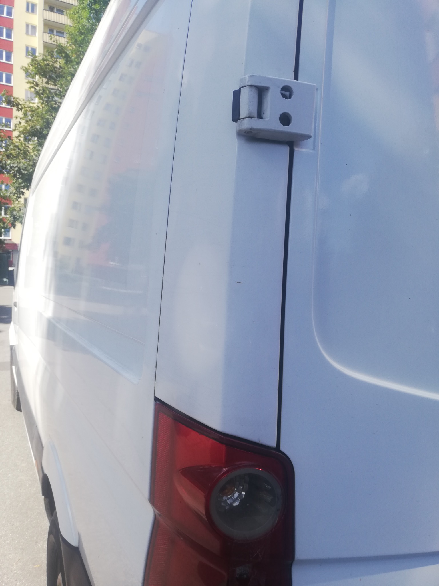 Zamknięte tylne drzwi białego busa z widocznym światłem stop i zawiasem, na tle bloku mieszkalnego.