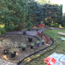 Przygotowanie terenu pod nowy ogród: wytyczona ścieżka z obrzeżami, rozłożona agrowłóknina, doniczki z roślinami, worki ziemi i narzędzia ogrodnicze w tle drzew