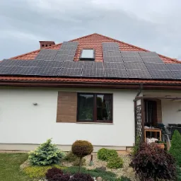 Dom jednorodzinny z czerwoną dachówką i panelami słonecznymi ułożonymi w trzech sekcjach, widok z poziomu ogrodu z dekoracyjnymi krzewami.