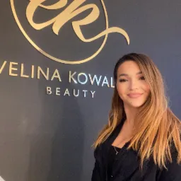 Portret kobiety z delikatnym makijażem, stojącej na tle ściany z logo 'Ewelina Kowal Beauty' w złotych literach.