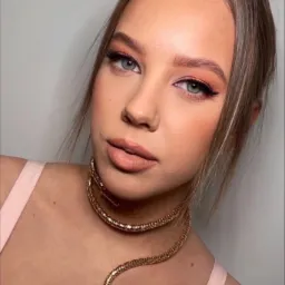 Portret kobiety z profesjonalnym makijażem oczu i ust w odcieniach brzoskwini, podkreślony złotym naszyjnikiem w kształcie węża. Subtelne konturowanie twarzy.