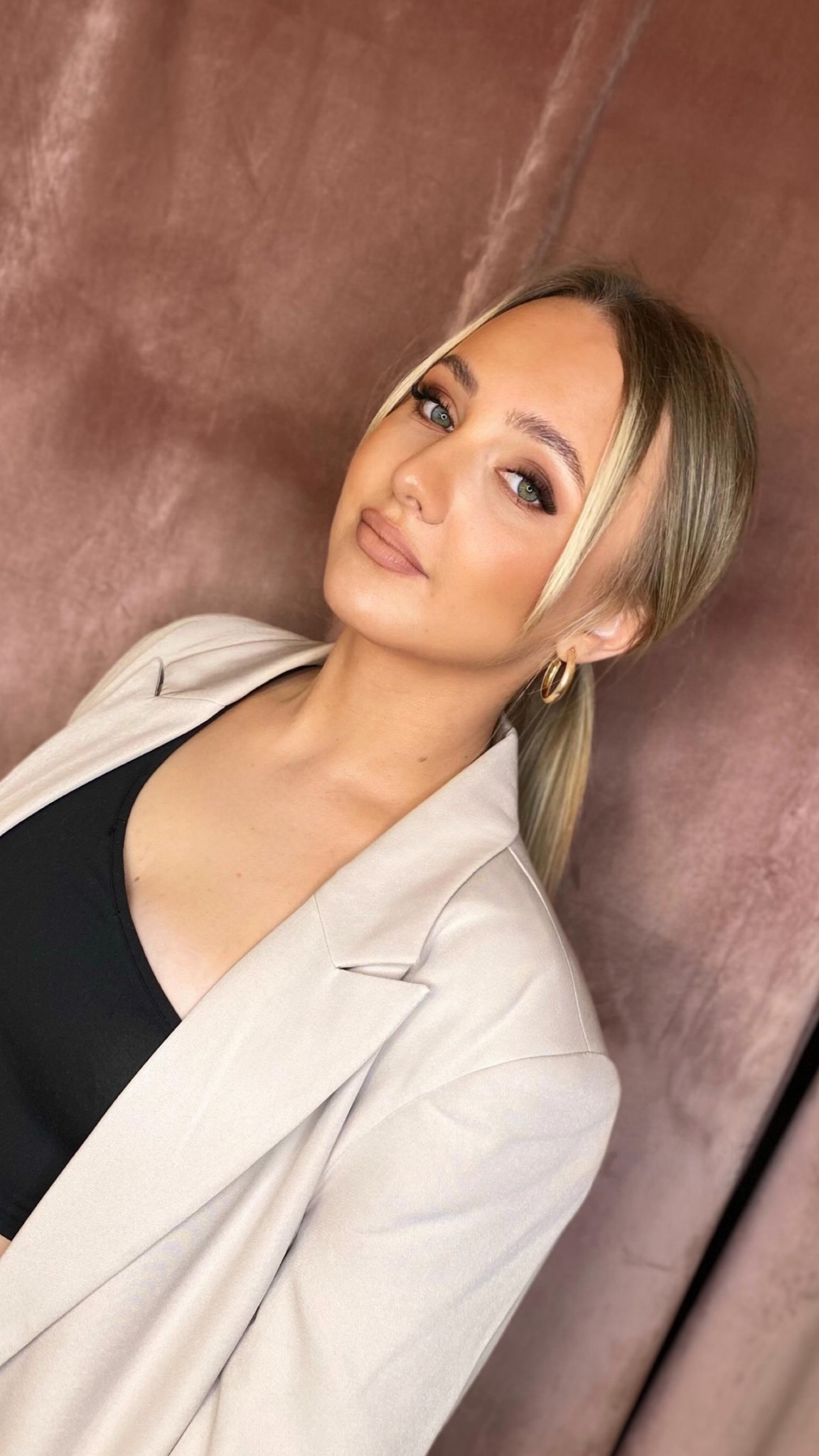 Portret kobiety z profesjonalnym makijażem, beżowy cień na powiekach, brzoskwiniowe usta, złote kolczyki koła, blond włosy z pasemkami i beżowa marynarka na tle pudrowego różu.