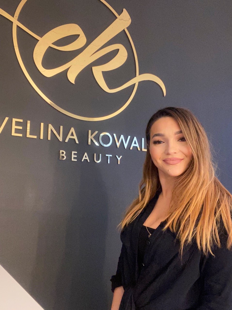 Portret kobiety z delikatnym makijażem, stojącej na tle ściany z logo 'Ewelina Kowal Beauty' w złotych literach.