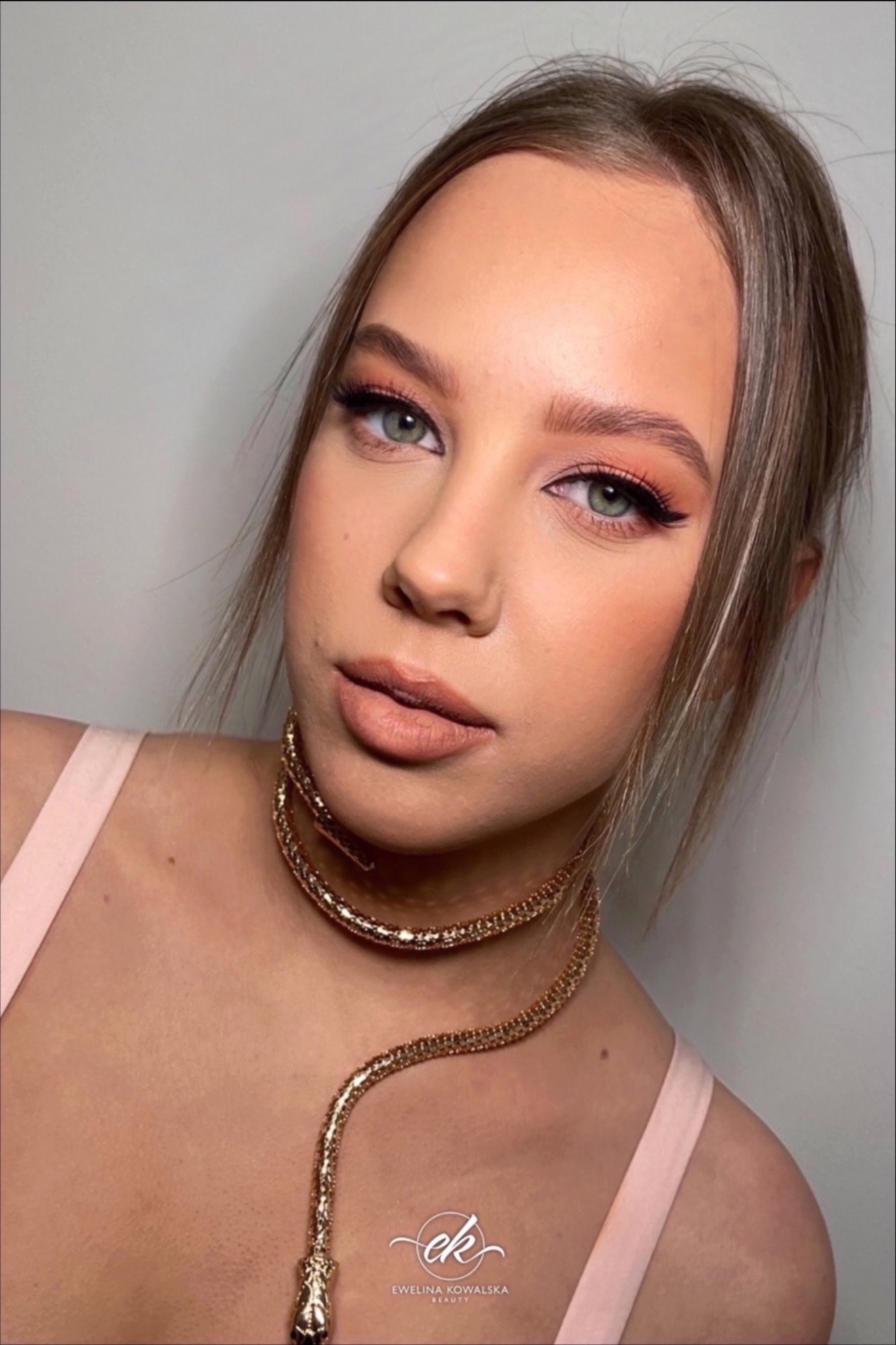 Portret kobiety z profesjonalnym makijażem oczu i ust w odcieniach brzoskwini, podkreślony złotym naszyjnikiem w kształcie węża. Subtelne konturowanie twarzy.