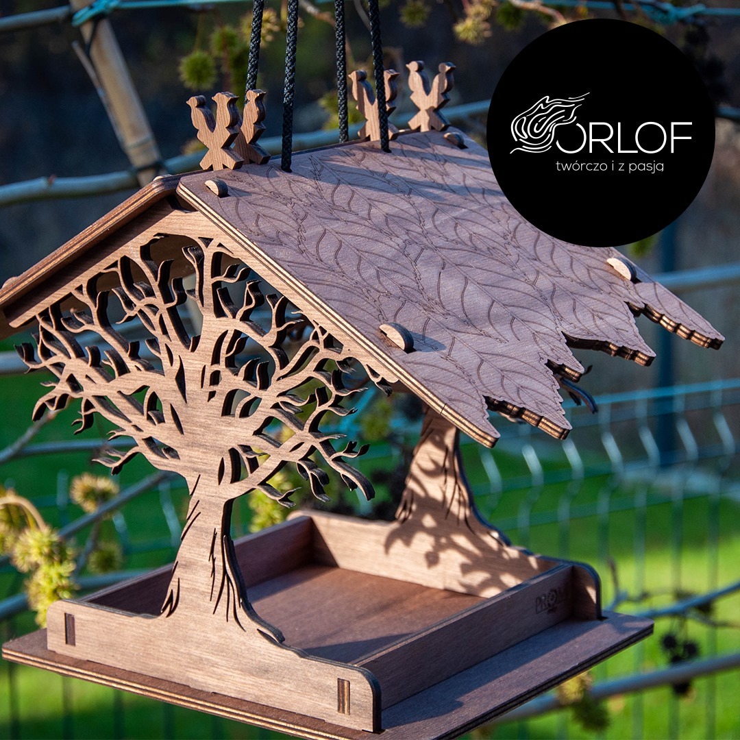 Drewniany karmnik dla ptaków z laserowo wycinanym motywem drzewa i liści na dachu, zawieszony na sznurkach, z logo firmy w prawym górnym rogu.