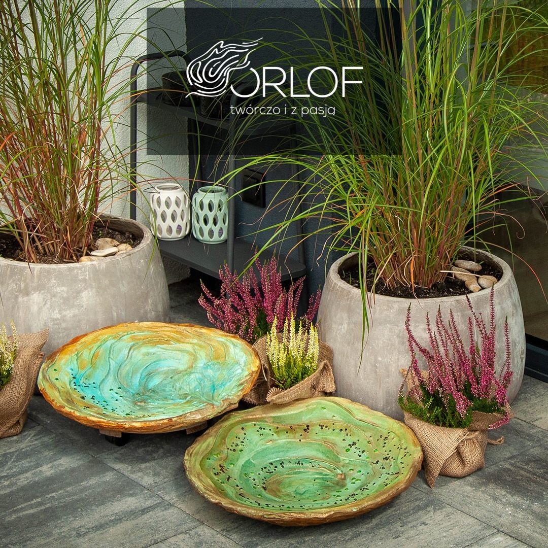 Kompozycja z doniczkowymi trawami ozdobnymi w betonowych donicach, ceramicznymi misami w odcieniach zieleni i błękitu oraz wrzosami w jutowych osłonkach, na tle szarej elewacji z logo firmy.