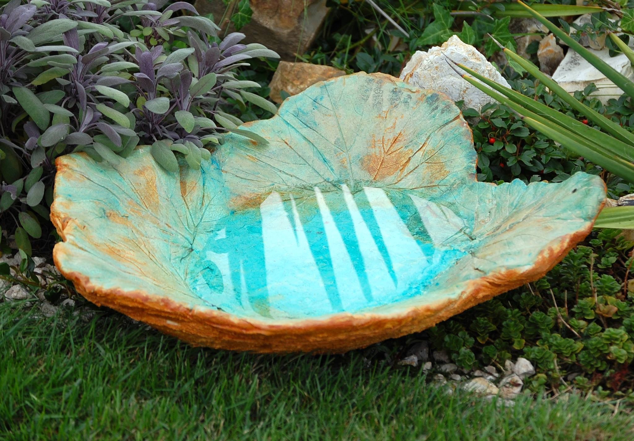Dekoracyjna, ceramiczna misa w kształcie liścia, wypełniona wodą, otoczona zielenią ogrodową.
