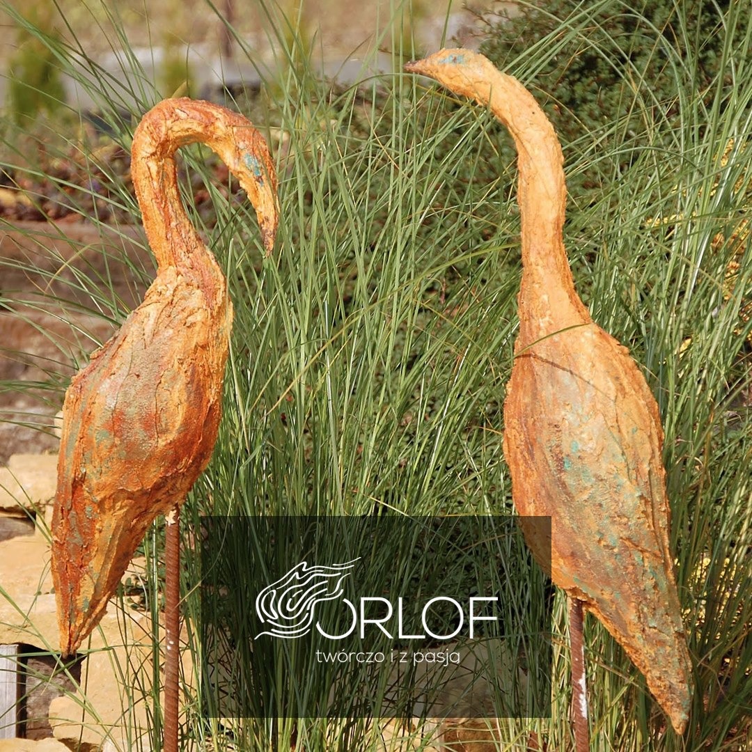 Dwie rzeźby ogrodowe przedstawiające stylizowane żurawie z pomarańczowo-turkusową patyną, umieszczone wśród zielonej trawy ozdobnej i kamiennej rabaty, z widocznym logo firmy ORLOF.