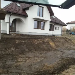 Przygotowany teren pod budowę domu jednorodzinnego z widocznym wykopem i elementami maszyny budowlanej w kadrze.