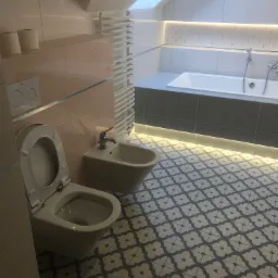 Wykończona łazienka na poddaszu z oknem dachowym, z toaletą, bidetem, grzejnikiem drabinkowym i wanną z podświetleniem LED, utrzymana w jasnej kolorystyce z wzorzystą podłogą.