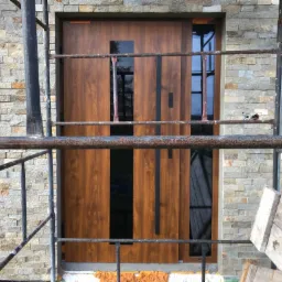 Brązowe drzwi wejściowe z czarnymi szybami i pionowymi elementami dekoracyjnymi, zamontowane w elewacji z kamienia naturalnego, widoczne zza rusztowania.