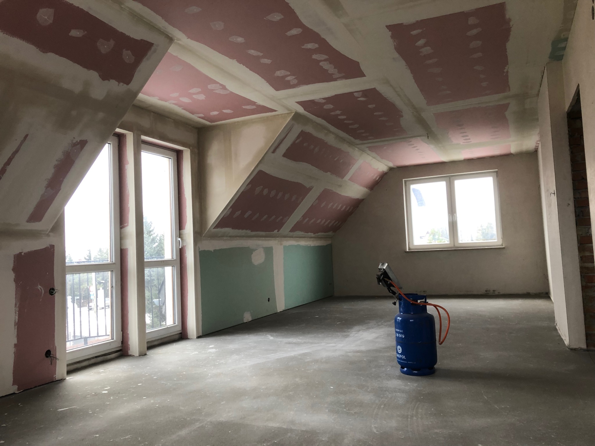 Poddasze w trakcie prac wykończeniowych: widoczne płyty kartonowo-gipsowe na skośnym suficie i ścianach, częściowo zaszpachlowane, betonowa wylewka na podłodze, okna z widokiem na zewnątrz...
