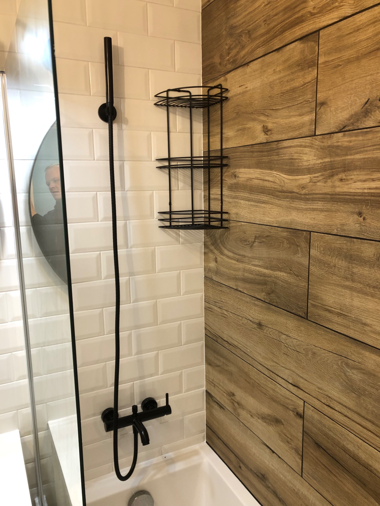 Nowoczesna łazienka z czarną armaturą, białą glazurą typu metro i ścianą wyłożoną płytkami imitującymi drewno, z widocznym odbiciem osoby w okrągłym lustrze.