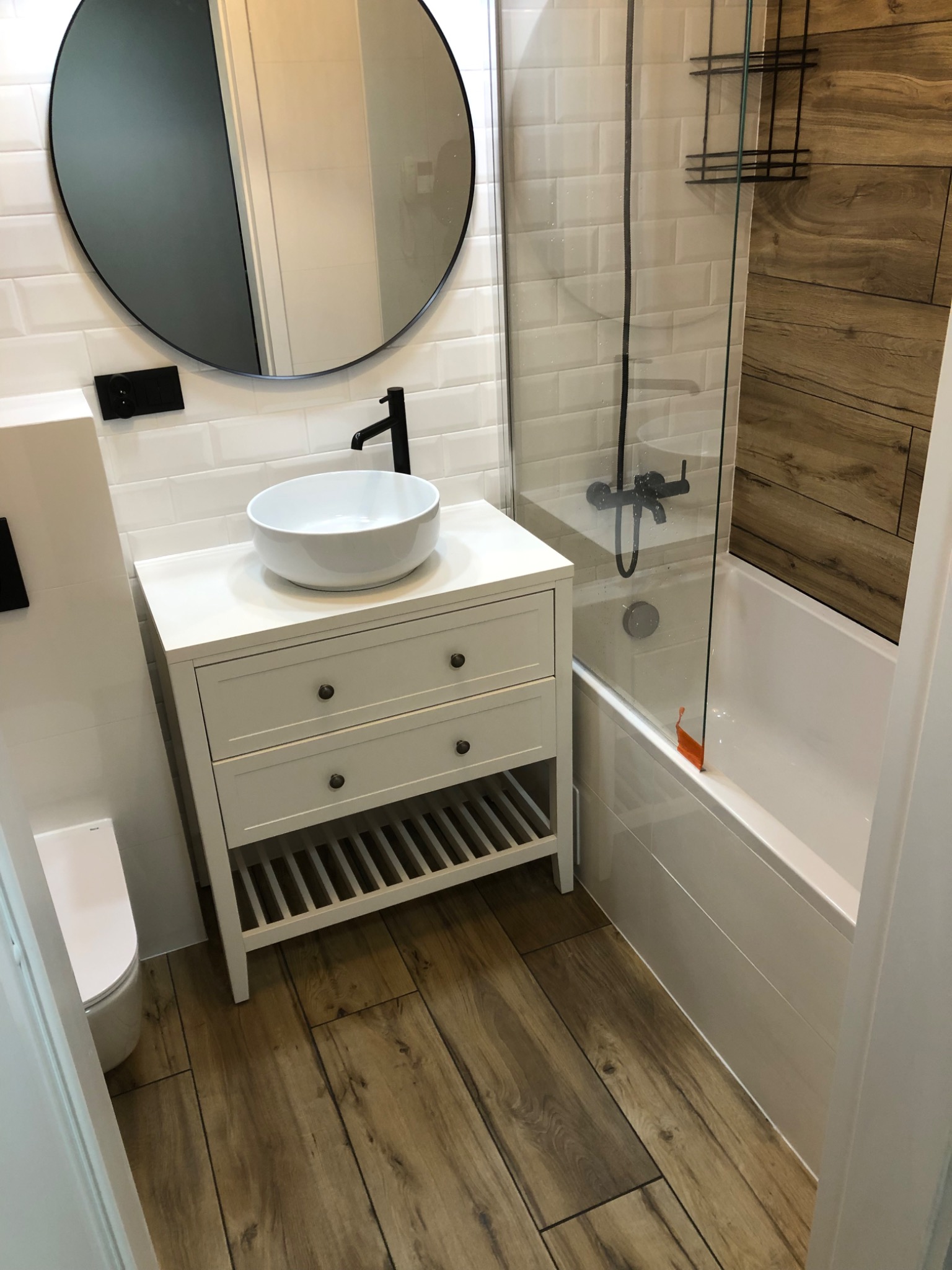 Nowoczesna łazienka z drewnopodobną podłogą, białą szafką z umywalką nablatową, okrągłym lustrem i kabiną prysznicową z drewnianym panelem bocznym.