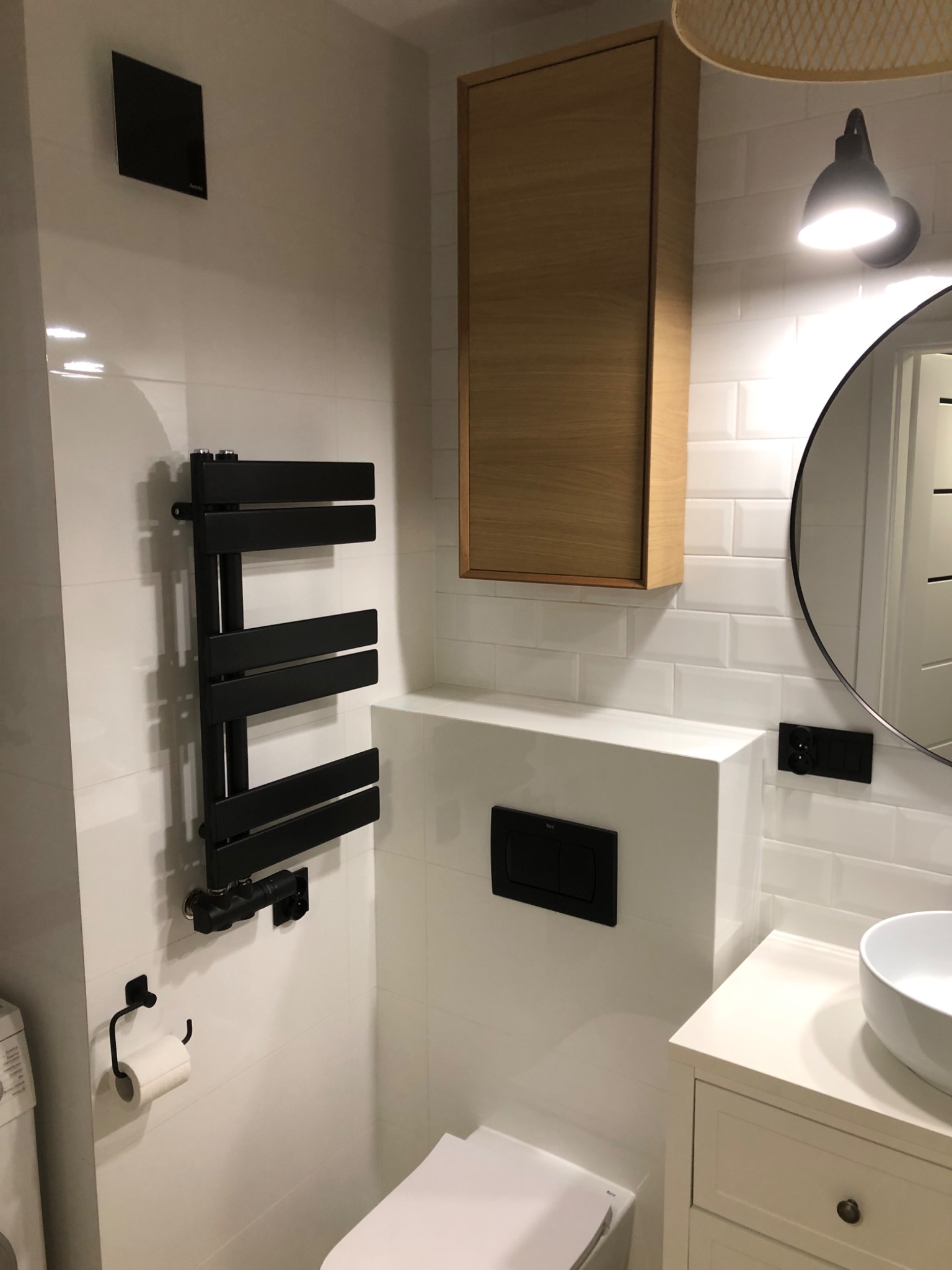 Nowoczesna łazienka z białymi płytkami typu metro, czarnym grzejnikiem drabinkowym, okrągłym lustrem i drewnianą szafką wiszącą, oświetlona punktową lampą.