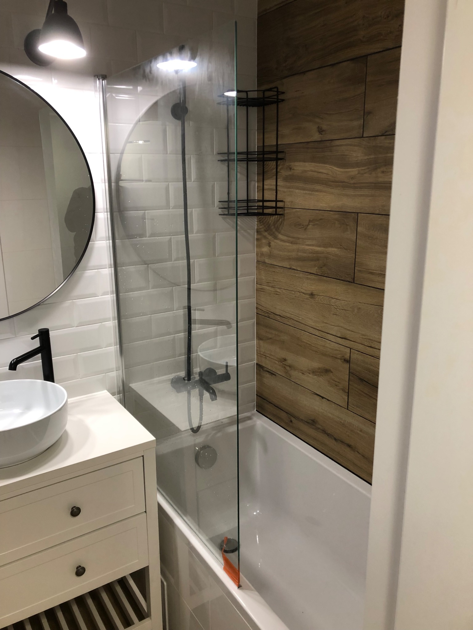 Nowoczesna łazienka z białymi płytkami metra, drewnopodobnymi panelami ściennymi, kabiną prysznicową ze szkła hartowanego i okrągłym lustrem z czarną ramą, czarna bateria umywalkowa