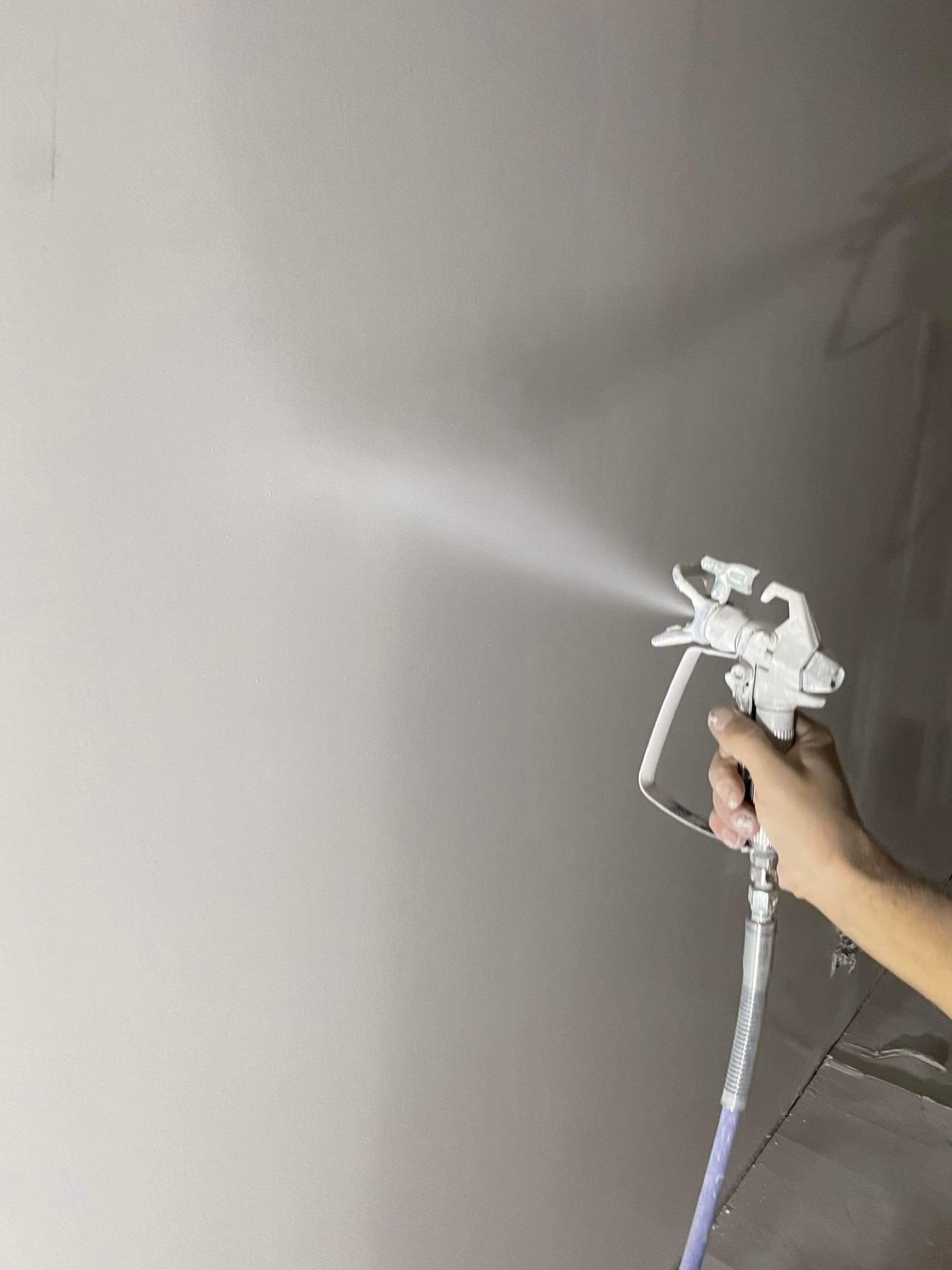 Malowanie ściany agregatem natryskowym w Kątach Wrocławskich. Widoczna ręka trzymająca pistolet natryskowy i rozpylana farba na gładkiej powierzchni.