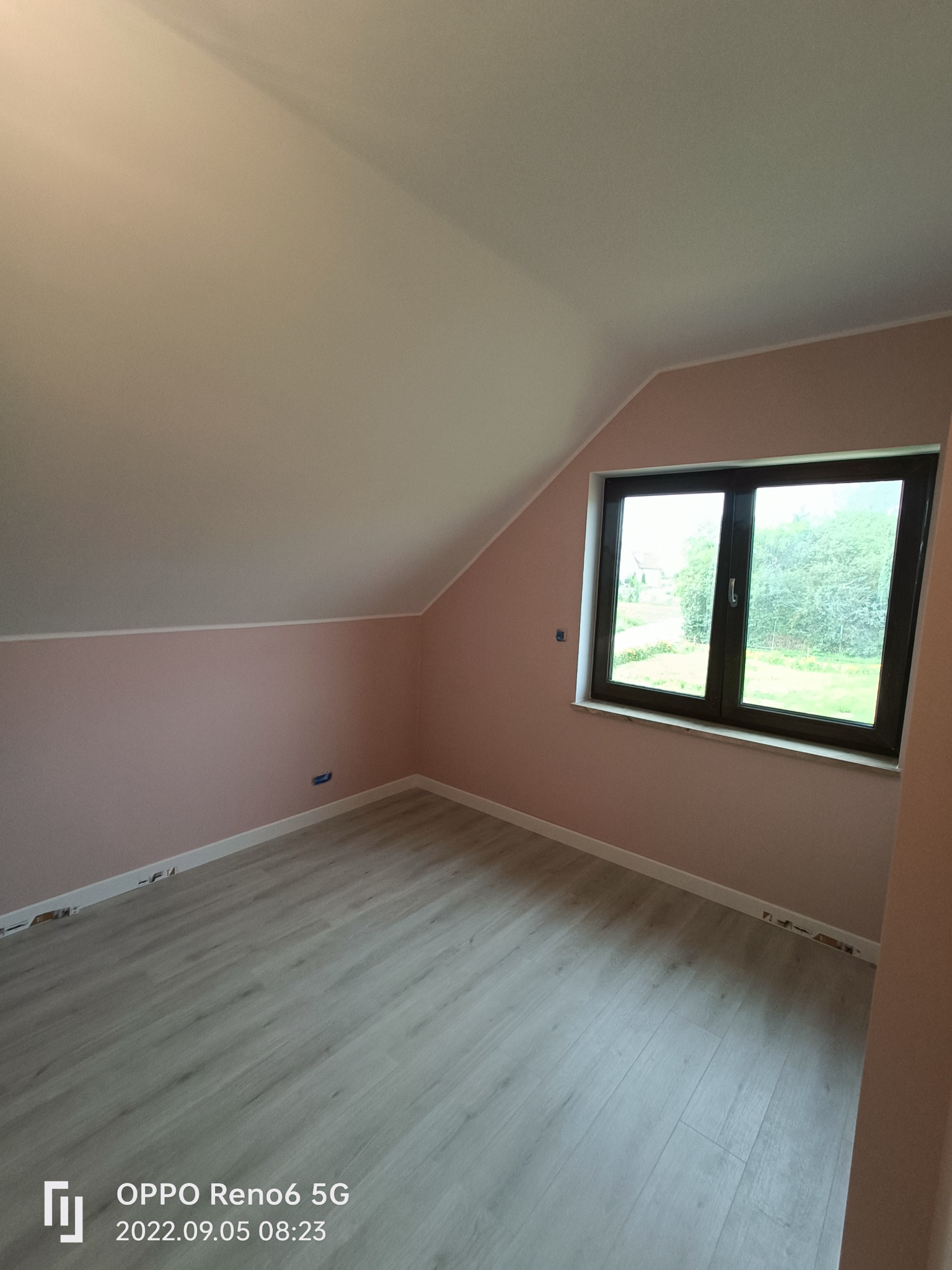 Pomalowane na różowo poddasze ze świeżo ułożonymi panelami podłogowymi, widok na zieleń przez okno.