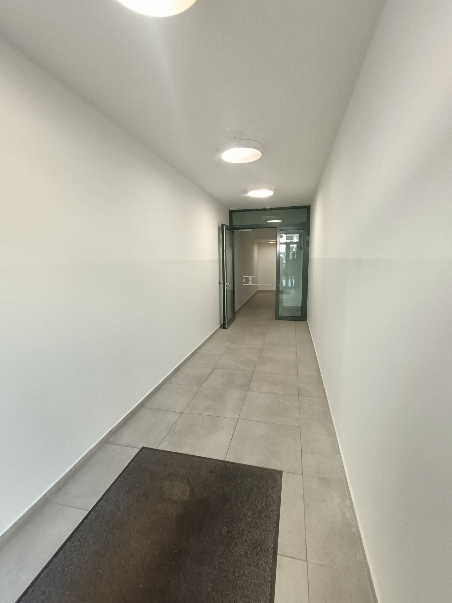 Korytarz biurowy po odświeżeniu w Kątach Wrocławskich, z jasnymi ścianami pomalowanymi do pewnej wysokości, szarą podłogą z płytek i wycieraczką przy wejściu.