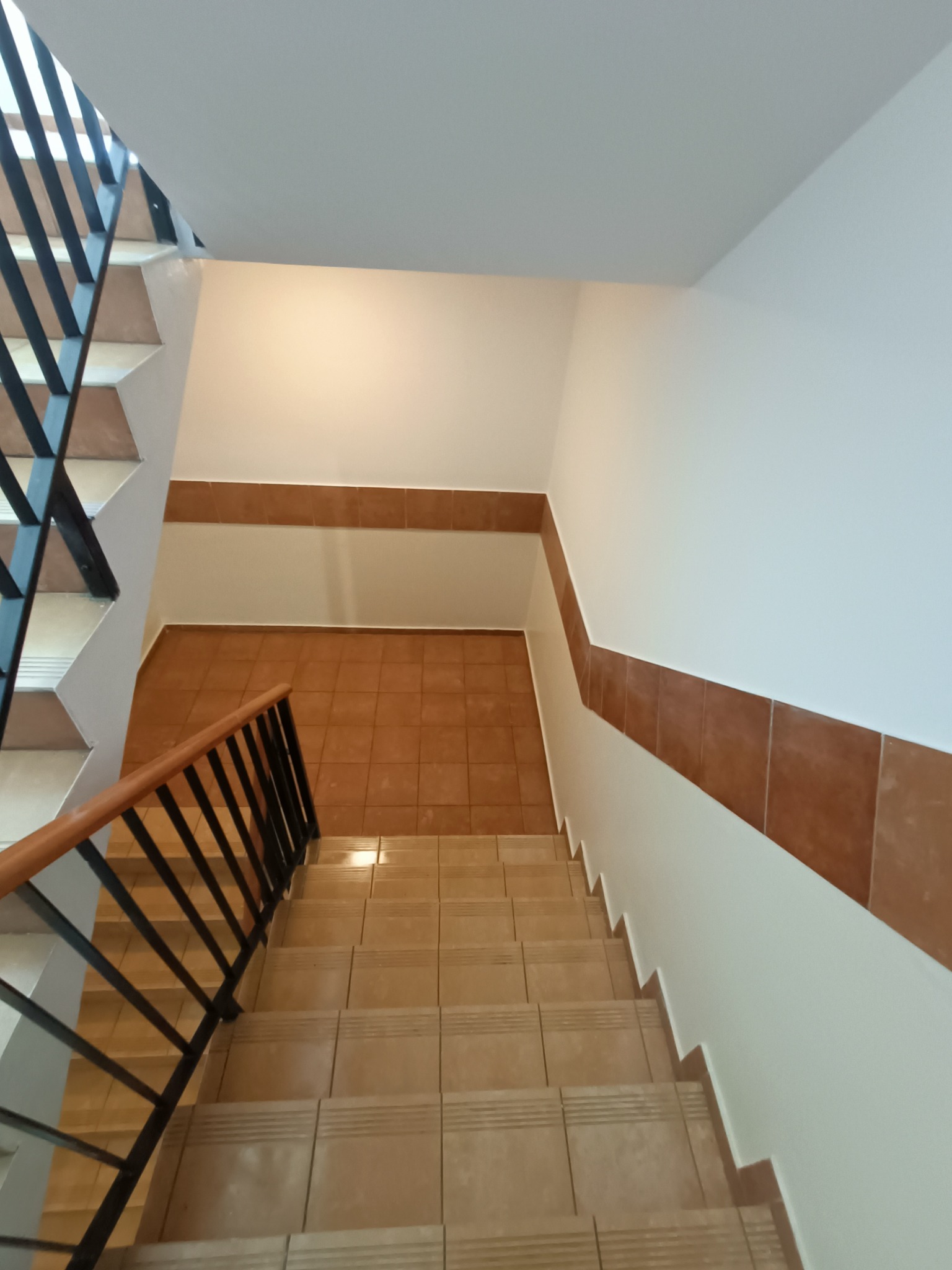 Klatka schodowa z ceramicznymi płytkami w odcieniach beżu i brązu, białe ściany z brązowym pasem płytek wzdłuż biegu schodów, czarna metalowa balustrada z drewnianym poręczą, widok z góry.