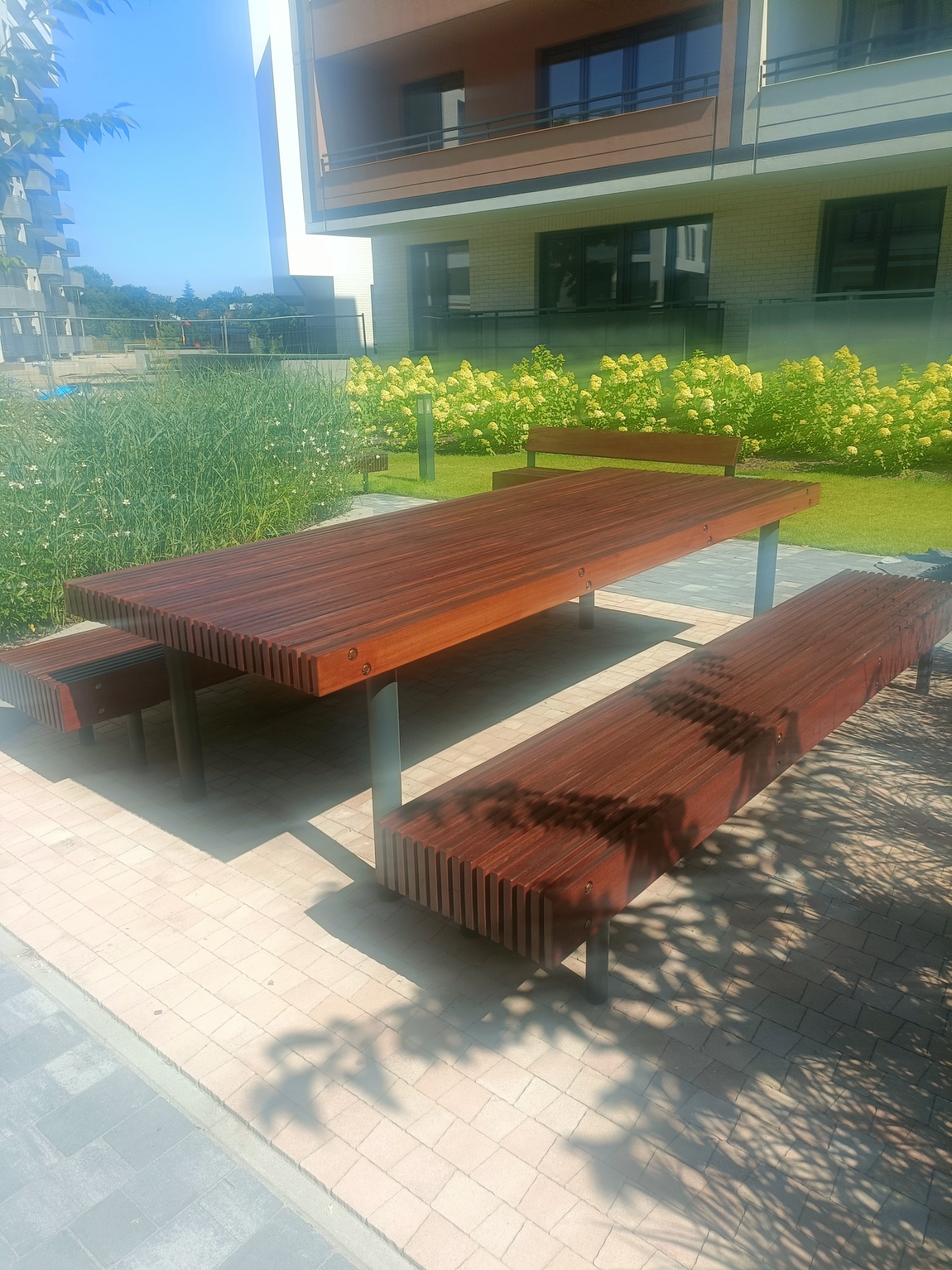 Drewniany stół piknikowy z ławkami na zewnątrz nowoczesnego budynku, oświetlony jasnym słońcem rzucającym cienie.