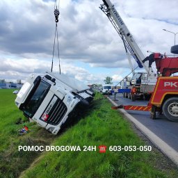 Auto-Lukas Pomoc Drogowa - Serwis Mobilny BUS-TIR 24H - Biała ciężarówka DAF po wypadku, leżąca na boku w trawie, podnoszona przez dźwig lawety PKT Kosłom, widoczni pracownicy w odblaskowych kamizelkach.