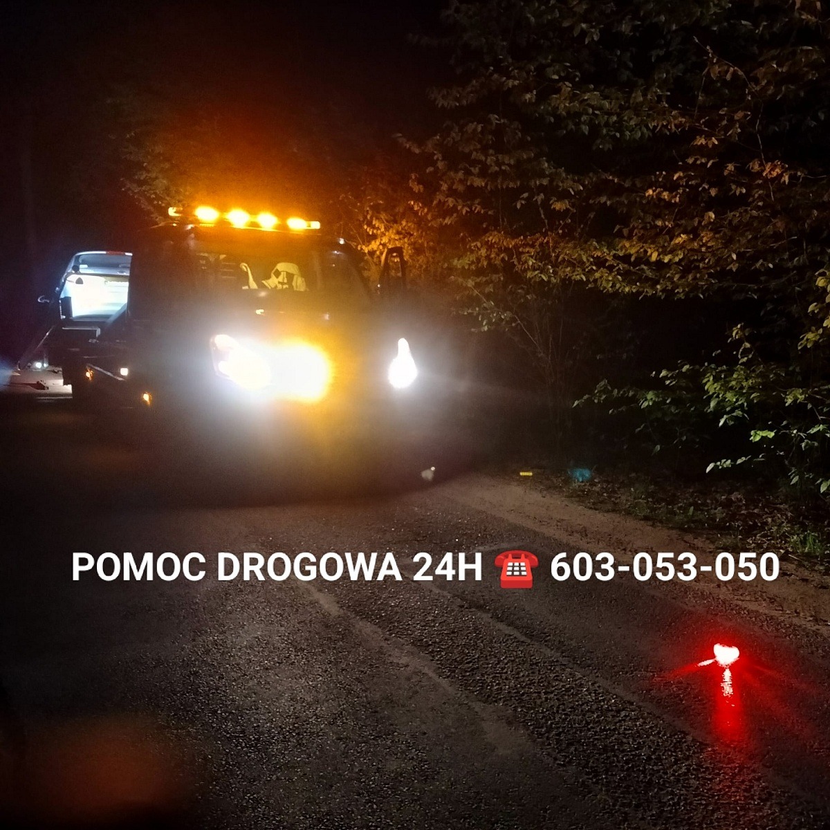 Laweta z włączonymi światłami awaryjnymi i przewożonym samochodem nocą na leśnej drodze, widoczny numer telefonu 'Pomoc Drogowa 24H'.