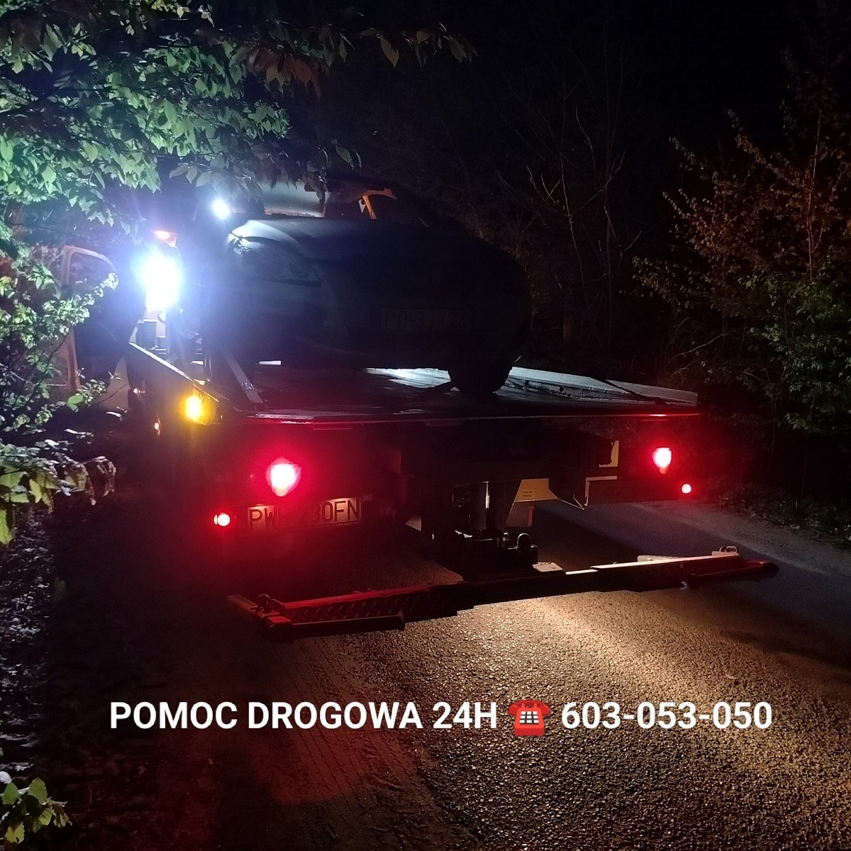 Samochód osobowy załadowany na platformę lawety holowniczej w nocy, w tle drzewa. Widoczne światła lawety i tablica rejestracyjna. Na dole zdjęcia napis POMOC DROGOWA 24H i numer telefonu.