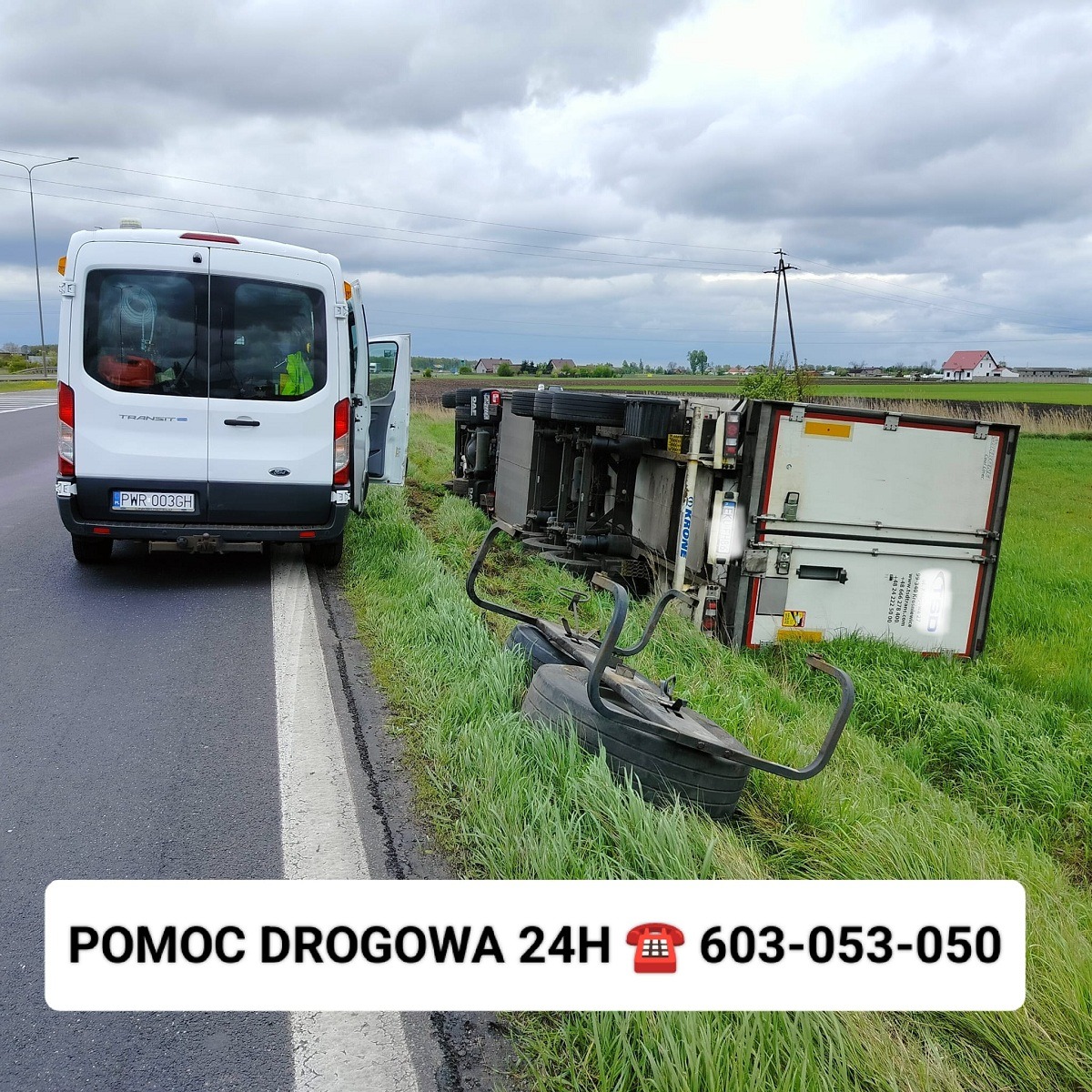 Przewrócona na bok naczepa ciężarówki w przydrożnym rowie, widoczny uszkodzony element podwozia, na asfalcie biały bus pomocy drogowej z otwartymi drzwiami.