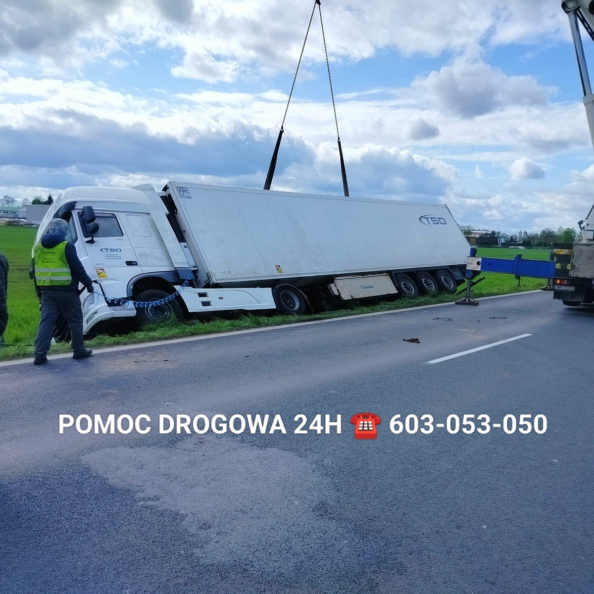 Uszkodzona ciężarówka z naczepą, częściowo podniesiona przez dźwig na poboczu drogi, widoczny pracownik w kamizelce odblaskowej, na asfalcie napis 'Pomoc Drogowa 24h' i numer telefonu.