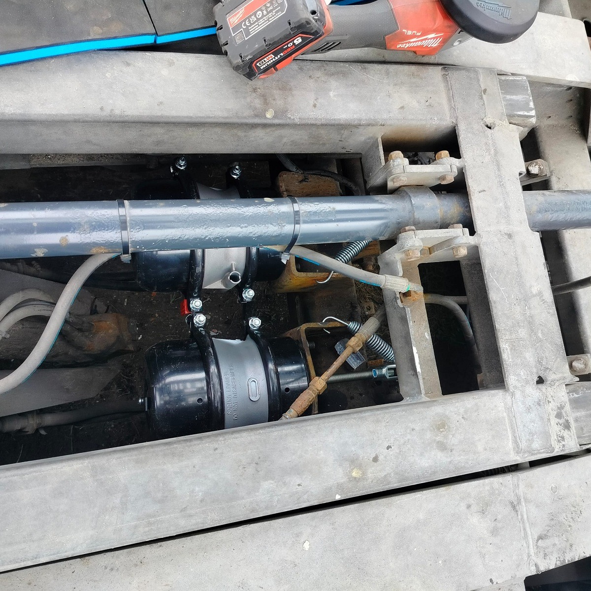 Widok z góry na zamontowane siłowniki pneumatyczne w metalowej ramie, widoczne przewody, śruby mocujące, oraz akumulator i szlifierka kątowa.