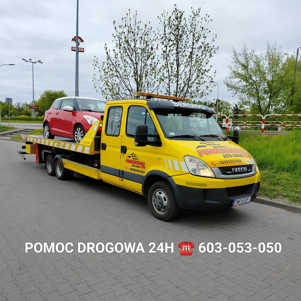 Żółta laweta z czerwonym samochodem osobowym na platformie, zaparkowana na poboczu drogi w pochmurny dzień, z widocznym logo firmy 'Pomoc Drogowa Auto-Lukas 24h' i numerem telefonu.