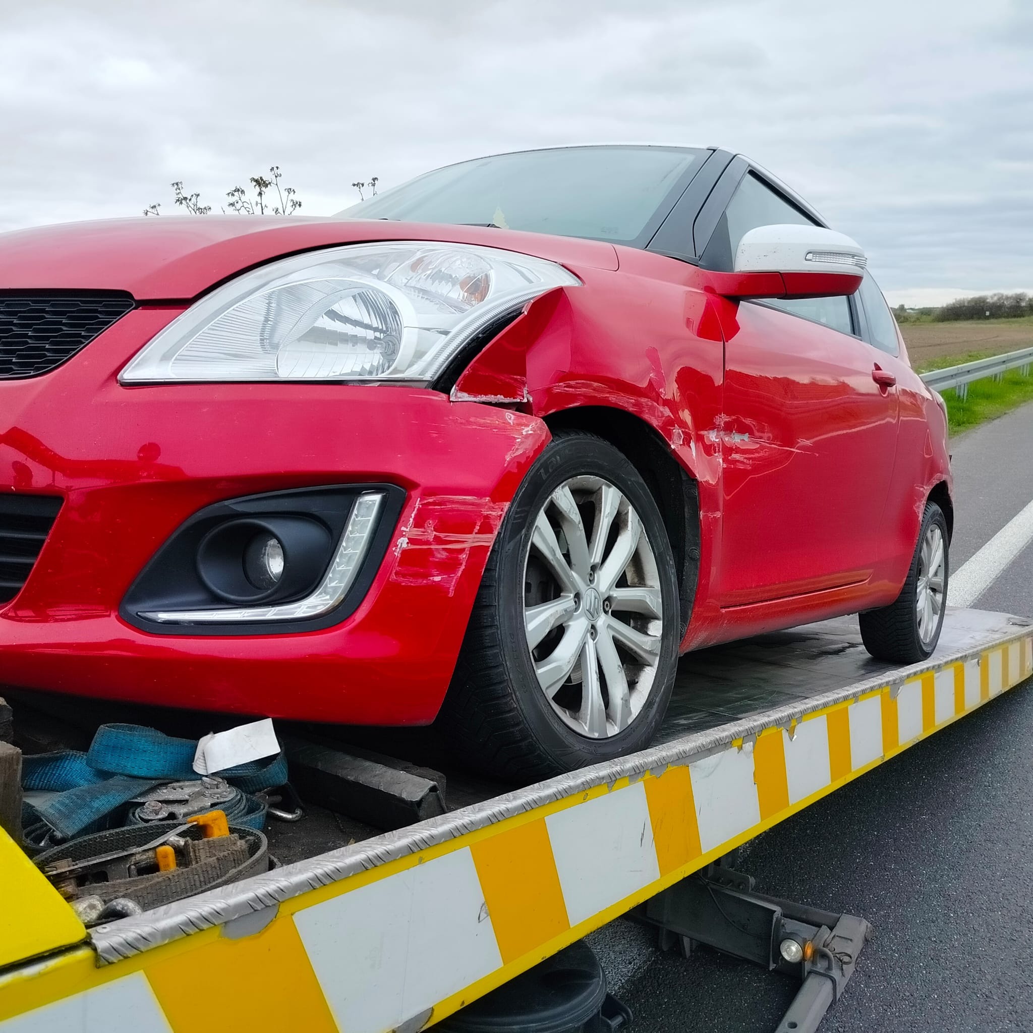 Czerwony samochód Suzuki Swift z uszkodzeniami karoserii, załadowany na lawetę z żółto-białymi pasami, widoczne niebieskie pasy mocujące, w tle szara nawierzchnia drogi i pochmurne niebo.
