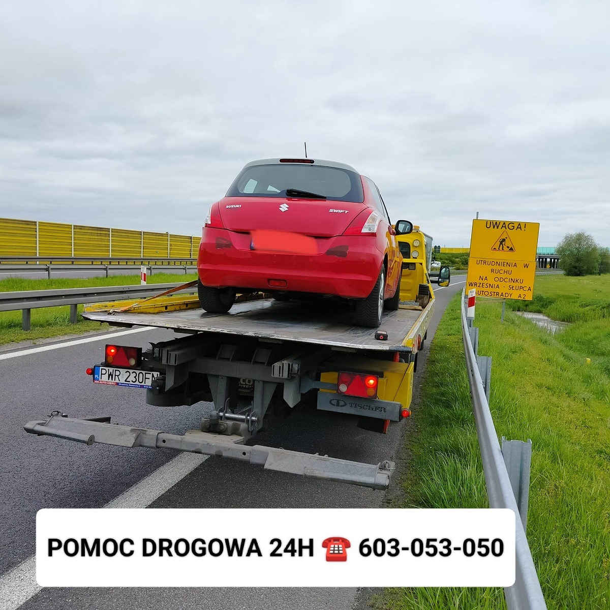 Czerwony samochód Suzuki Swift przewożony na żółtej lawecie na autostradzie A2, widoczny znak ostrzegawczy o utrudnieniach w ruchu.