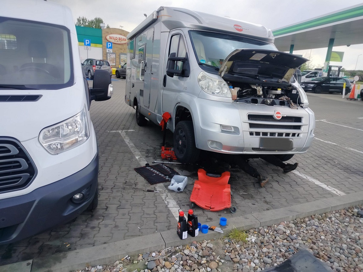 Srebrny kamper marki Burstner z otwartą maską, pod nim widoczne nogi osoby naprawiającej, obok rozłożone narzędzia, lewarek, kanisterki z olejem, a w tle biały bus i stacja benzynowa.