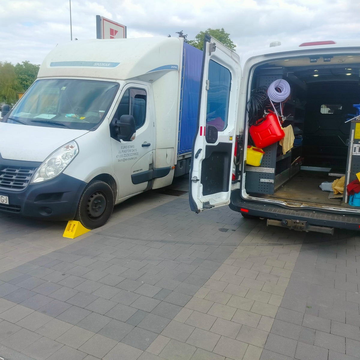 Dwa białe busy, jeden z plandeką i żółtymi klinami pod kołami, drugi z otwartymi tylnymi drzwiami i widocznym wyposażeniem warsztatowym wewnątrz, stoją na brukowanym placu.