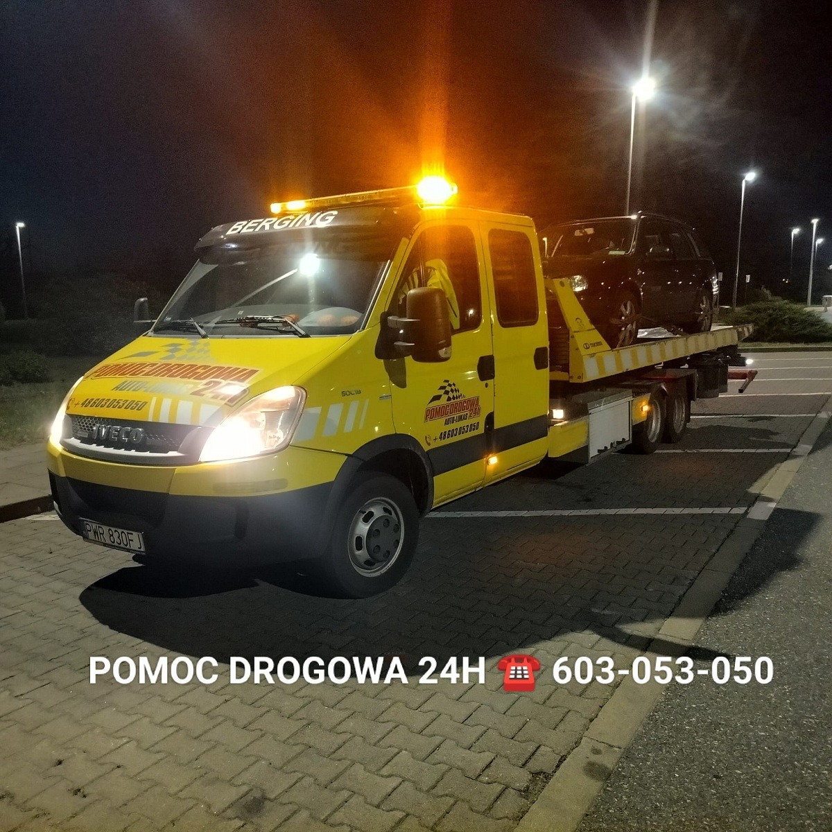 Żółta laweta Iveco z włączonymi światłami ostrzegawczymi przewozi czarny samochód osobowy na platformie w nocy, widoczne logo firmy 'POMOC DROGOWA' i numer telefonu alarmowego.
