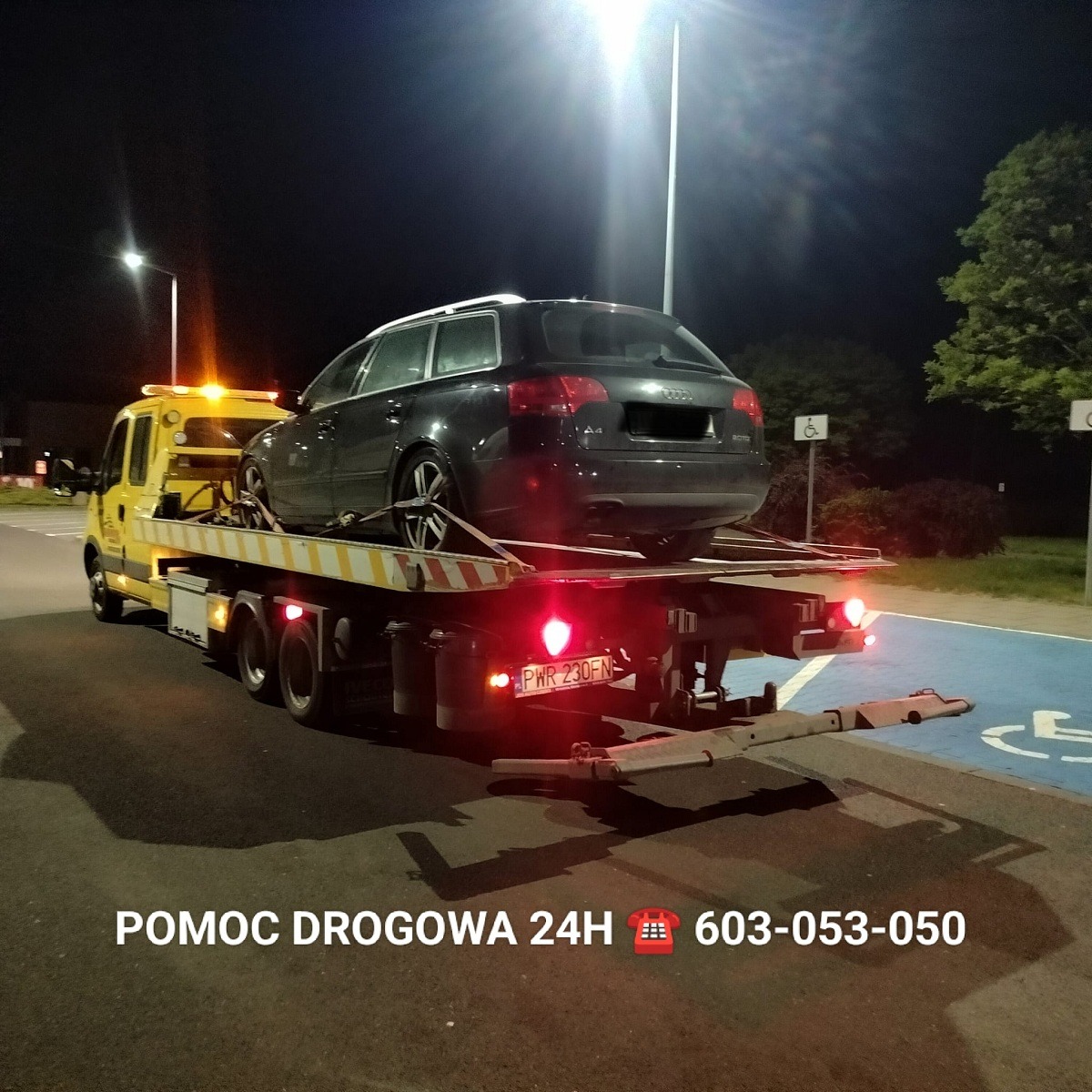 Ciemny samochód osobowy marki Audi A4 kombi z uszkodzonym kołem, zabezpieczony pasami transportowymi, przewożony na żółtej lawecie w nocy na ulicy we Wrześni. W tle widoczne oświetlenie uliczne...