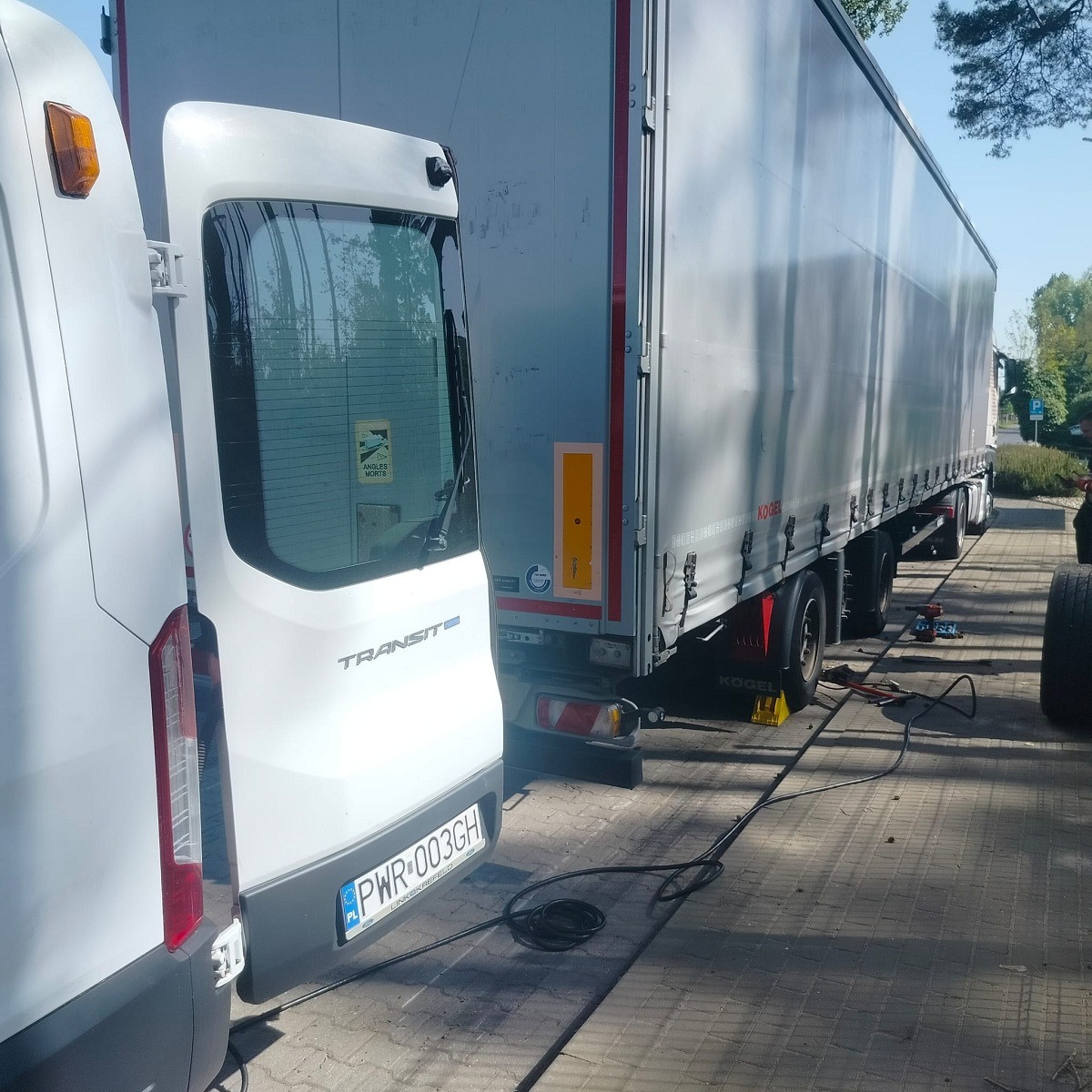 Biały bus Ford Transit i szara naczepa ciężarówki Kogel stoją obok siebie na kostce brukowej. Widoczne kable i narzędzia leżące na ziemi, sugerujące naprawę lub serwisowanie.