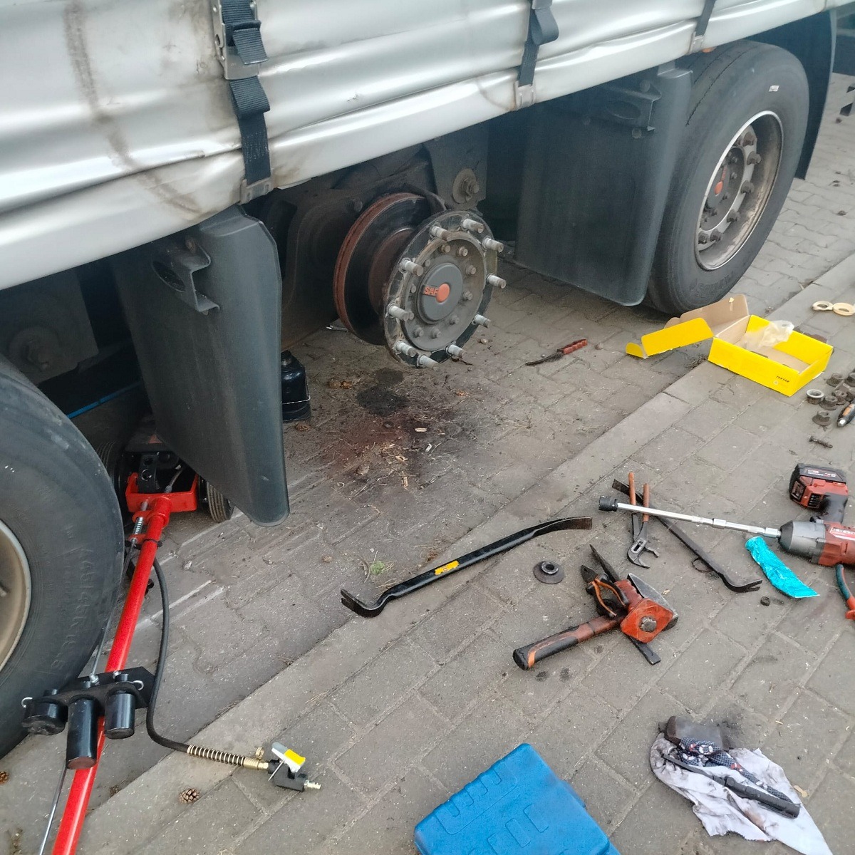 Ciężarówka z podnośnikiem hydraulicznym i zdjętym kołem, widoczne narzędzia i części zamienne na brukowanej nawierzchni.