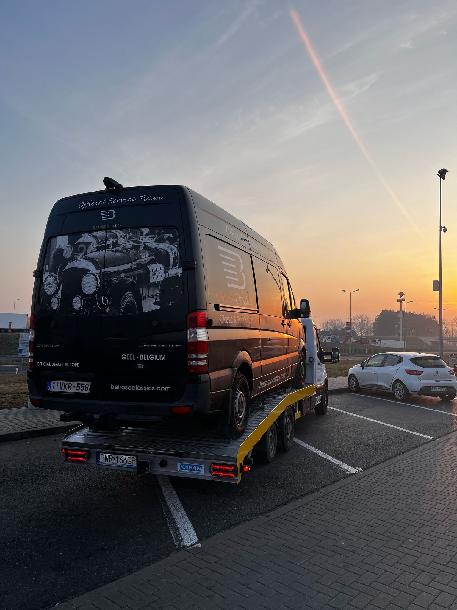 Czarna laweta marki Mercedes-Benz Sprinter z grafiką klasycznych aut na tylnych drzwiach, zaparkowana na parkingu o zmierzchu, z załadowanym na platformie szarym samochodem osobowym. Na tylnej...