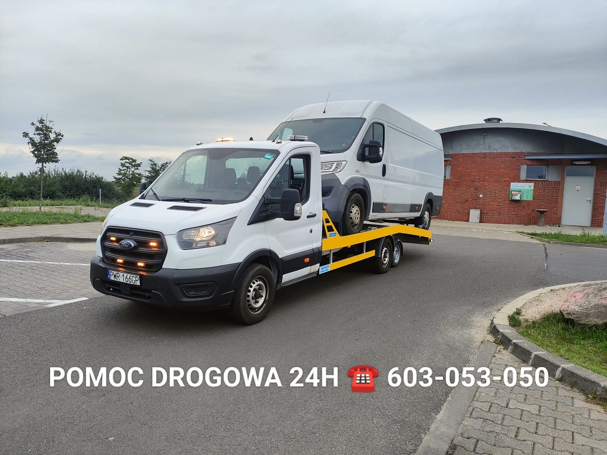 Biała laweta Ford z pomarańczowymi światłami przewozi białego busa na żółtej platformie. W tle budynek z czerwonej cegły i szare niebo.