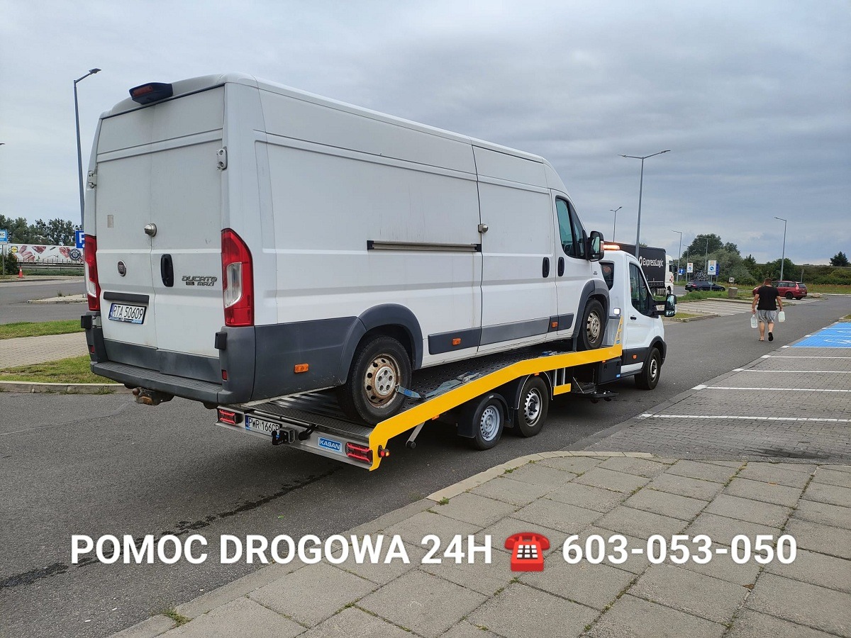 Biały bus na żółtej lawecie, zaparkowany na parkingu. Widoczny fragment chodnika i osoba w oddali. Pomoc drogowa z numerem telefonu na dole zdjęcia.